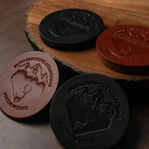 A set of four 4in round Chosen Leatherwork FAITH CAN MOVE MOUNTAINS Stamped English Bridle Leather coasters in black and brown, each embossed with a mountain scene, a deer, and the text FAITH CAN MOVE MOUNTAINS MATTHEW 17:20.