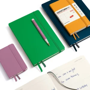 A green Leuchtturm1917 2026 Weekly Planner Medium (A5), Hardcover, English with a pen resting on top, placed next to a small purple notebook, a large blue planner, and an open page showing handwritten schedule entries for June 2026.