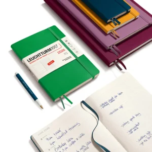A green Leuchtturm1917 2026 Weekly Planner & Ruled Notebook Composition (B5), Hardcover, displayed with a pen and a stack of colorful notebooks, one open to a page of handwritten notes.